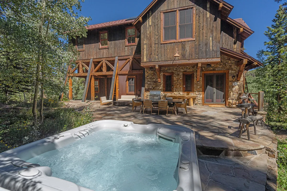 This inviting exterior image features a private hot tub on a wooden deck, surrounded by a stone wall and an outdoor dining area. The space is set against a backdrop of tranquil nature, providing a perfect setting for relaxing under the stars or gathering with friends and family for shared meals and conversation.