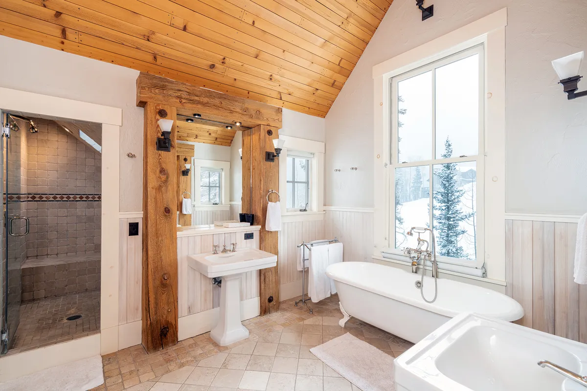 A serene bathroom featuring a freestanding clawfoot bathtub positioned next to large windows with views of a snowy landscape, rustic wooden beams overhead, and tranquil natural light filling the space.