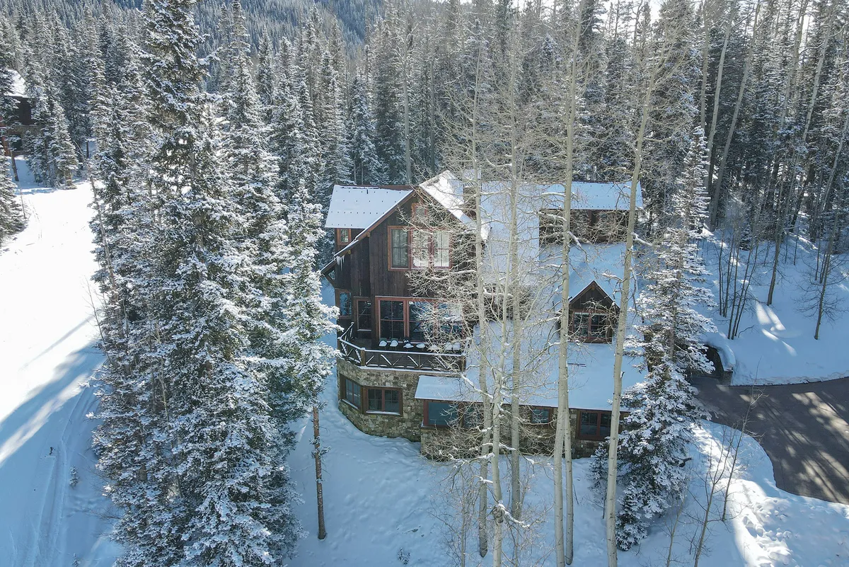 A charming, multi-level lodge built of wood and stone is surrounded by snow-laden trees. Balconies offer views of the forest, and the overall scene radiates peacefulness and privacy, connecting the home directly to its winter wonderland setting.
