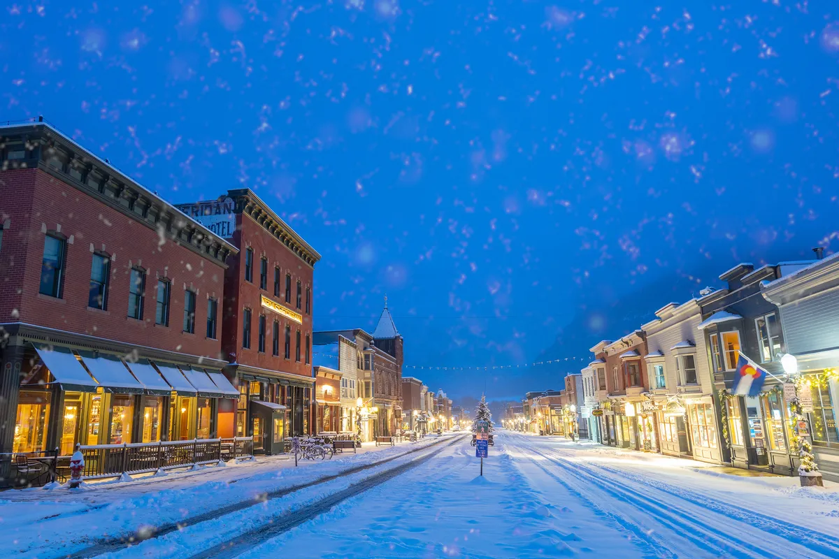 A charming, snow-covered street in Telluride, lined with decorated storefronts, and a brightly lit Christmas tree visible in the distance. The festive scene is bustling with the energy of holiday shopping and winter strolls.