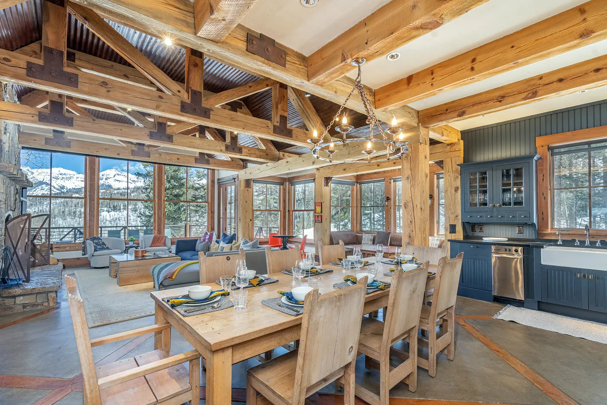 A spacious open-concept area combining the dining table, kitchen with modern appliances, a cozy fireplace, and comfortable seating, all set against a backdrop of large windows that let in natural light and reveal mountain views. The space is designed for group gatherings, meals, and relaxing by the fire after a day outdoors.
