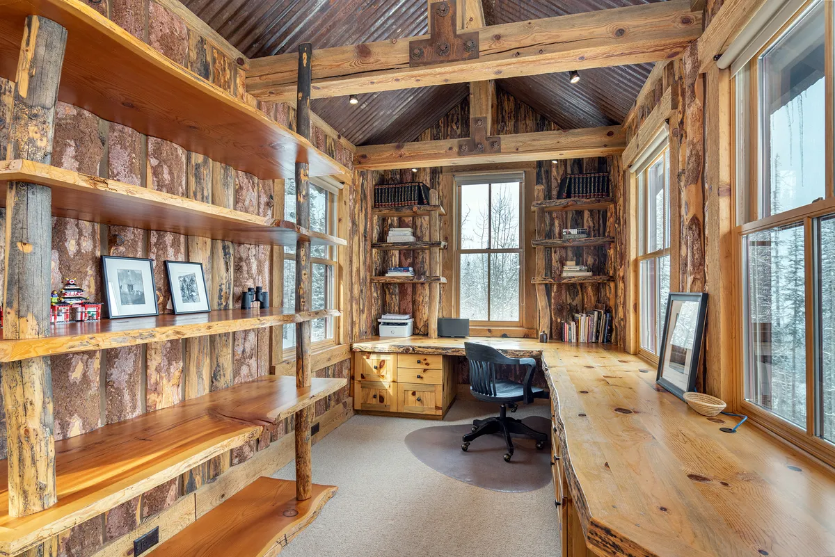 The image shows a cozy, rustic office space featuring wooden shelves, a large desk, and windows that frame inspiring mountain views. Natural light pours in, highlighting the inviting workspace that is perfect for focused work sessions and moments of tranquility.