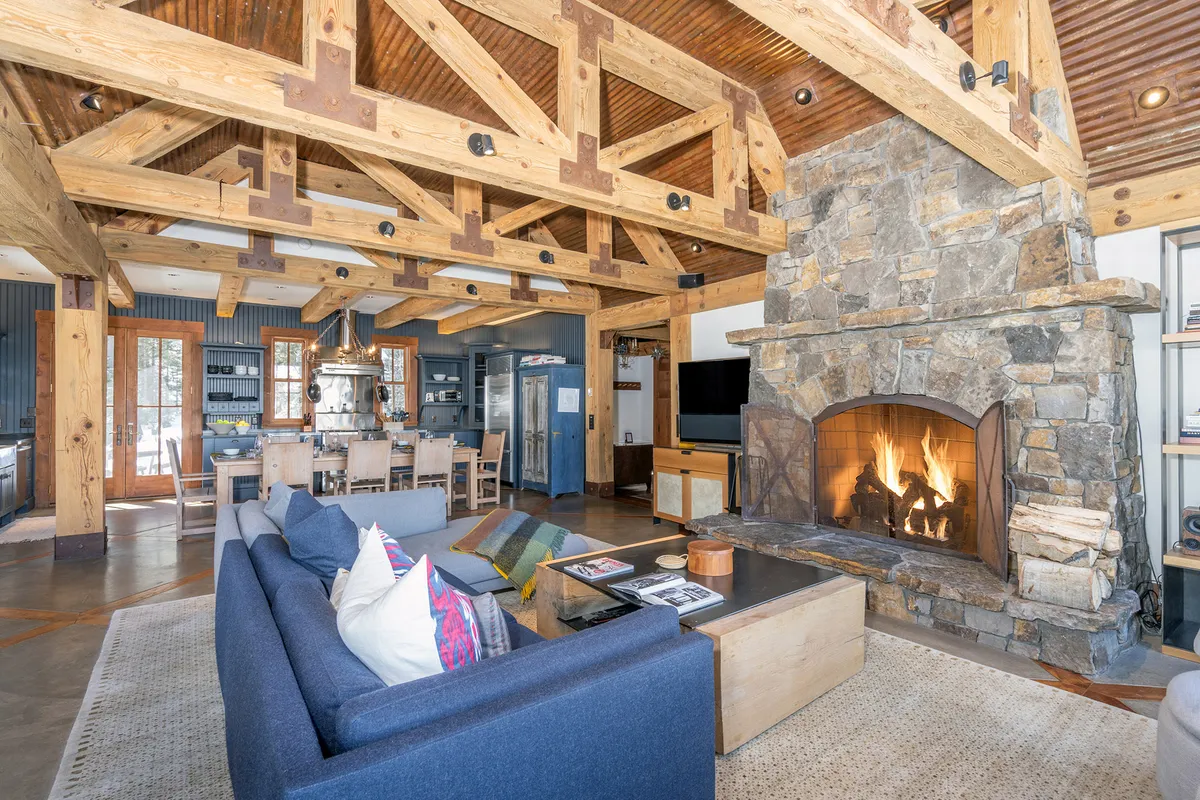 A warm, inviting living area featuring a striking stone fireplace, wooden beams overhead, a comfortable sofa with colorful pillows, and an adjacent dining area. The ambiance is perfect for fireside chats, family meals, and group activities.