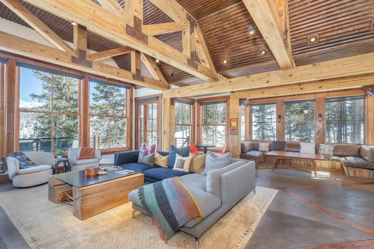 An expansive interior living area with a large sectional sofa, wooden beams, and floor-to-ceiling windows framing scenic sunset views. The welcoming atmosphere is perfect for group gatherings, game nights, or cozy fireside chats after a day outdoors.