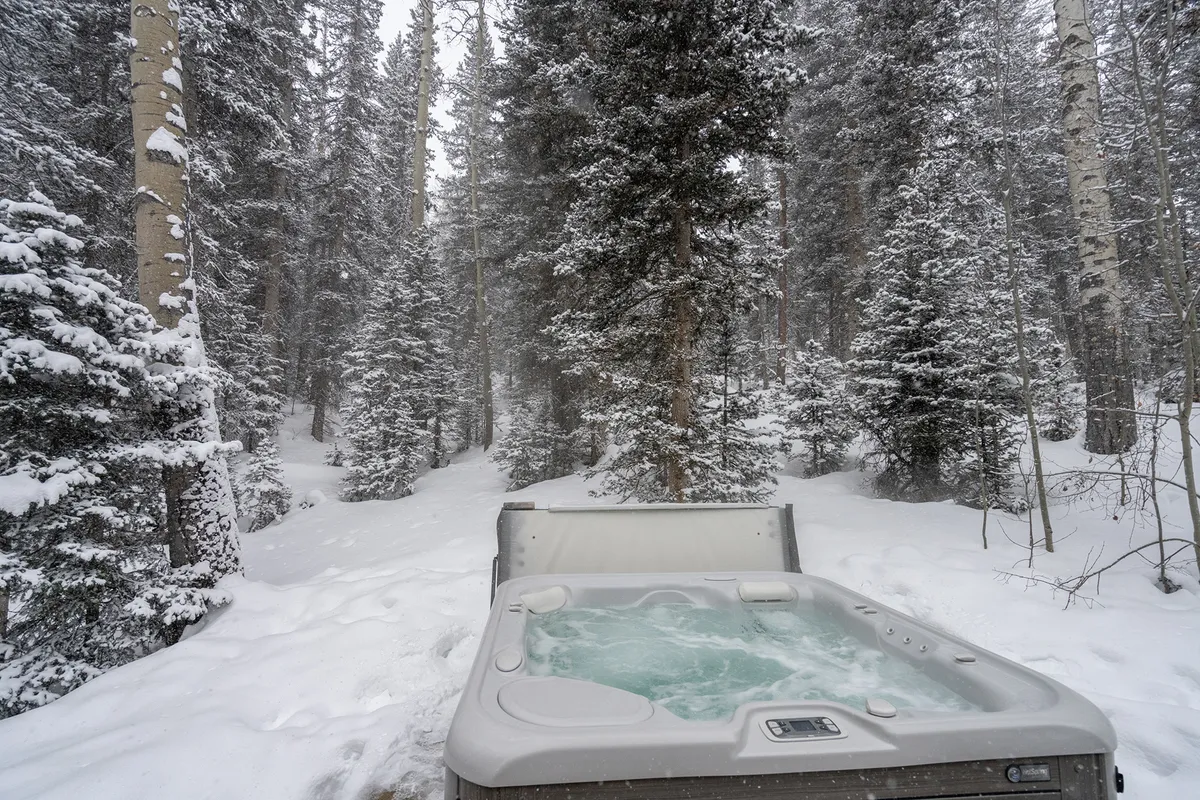 A private hot tub sits nestled among snow-covered trees and a forest pathway, creating a tranquil winter retreat. The steaming water contrasts beautifully with the surrounding white landscape, inviting relaxation after a day on the mountain.