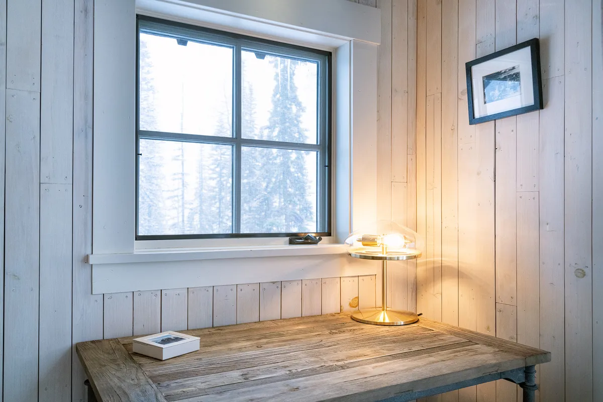 A calm, focused workspace with a wooden desk, lamp, and a window that overlooks natural scenery. The minimalistic setup exudes a peaceful atmosphere, ideal for reading, writing, or working in a serene environment.