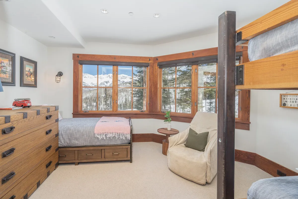 A welcoming bunk room with two bunk beds, a comfortable armchair beside a window, a wooden dresser, and expansive mountain views beyond the glass. The space radiates warmth and is perfect for children or teens.