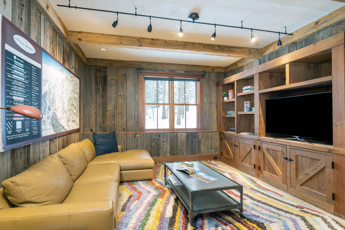 A cozy interior scene featuring a leather sectional sofa positioned on a colorful rug, with wooden walls and a television unit in the background. The arrangement invites group gatherings, movie nights, and relaxed socializing, all in a warm, welcoming atmosphere.