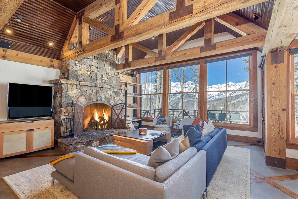 The image showcases a spacious, inviting living room with a grand stone fireplace at its center, surrounded by plush seating. Floor-to-ceiling windows frame breathtaking mountain views, filling the room with natural light and connecting the cozy interior with the dramatic Colorado landscape. The scene suggests relaxation by the fire, socializing, and soaking in the sunset vistas.
