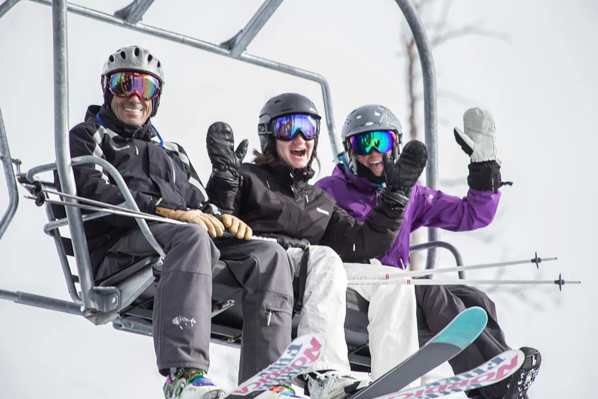 Three skiers riding a chairlift with snowy mountain peaks in the background, surrounded by a pristine winter landscape. Ski equipment and bundled attire emphasize the recreational spirit.