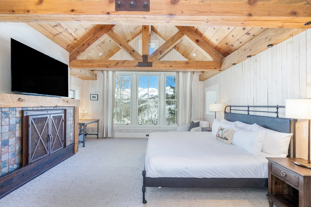 The image features a cozy bedroom with a rustic wooden beam ceiling, large windows showcasing breathtaking mountain views, a plush bed adorned with decorative pillows, and a fireplace adding warmth and ambiance.