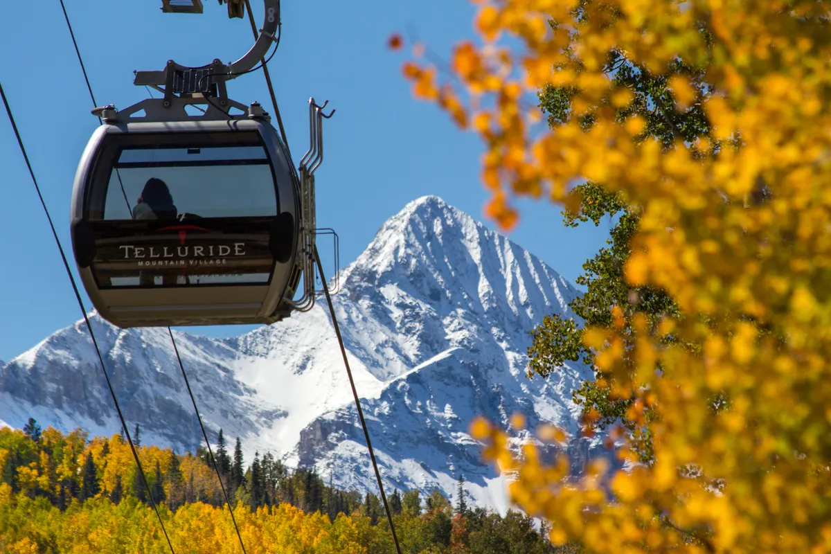 A dramatic exterior shot featuring a gondola soaring above vibrant autumn foliage, with snow-capped mountains in the distance. The image encapsulates the adventure and accessibility of mountain living, showing both natural beauty and a key local amenity.