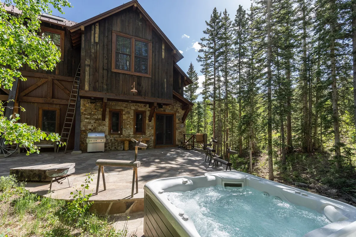 An exterior view of a bubbling hot tub on a wooden deck, surrounded by tall pine trees and a stone facade. The scene exudes tranquility and is perfect for stargazing, socializing, or unwinding after outdoor adventures in every season.