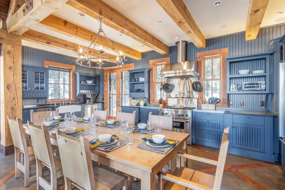 A large dining table sits beside a modern kitchen equipped with stainless steel appliances, blue cabinetry, and wooden beams, creating a welcoming space for group gatherings and culinary adventures.